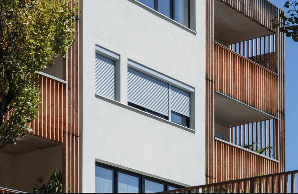 Moderní bytový dům s předokenními roletami v bílé barvě; elegantní kombinace s dřevěným lamelovým obkladem lodžií pro účinné stínění a soukromí.