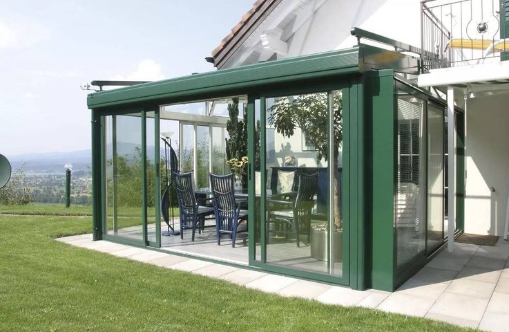 Moderní celoroční zimní zahrada v zelené hliníkové konstrukci s velkými posuvnými skleněnými stěnami vytváří u rodinného domu pohodlné místo pro posezení s výhledem do zahrady a okolní krajiny.