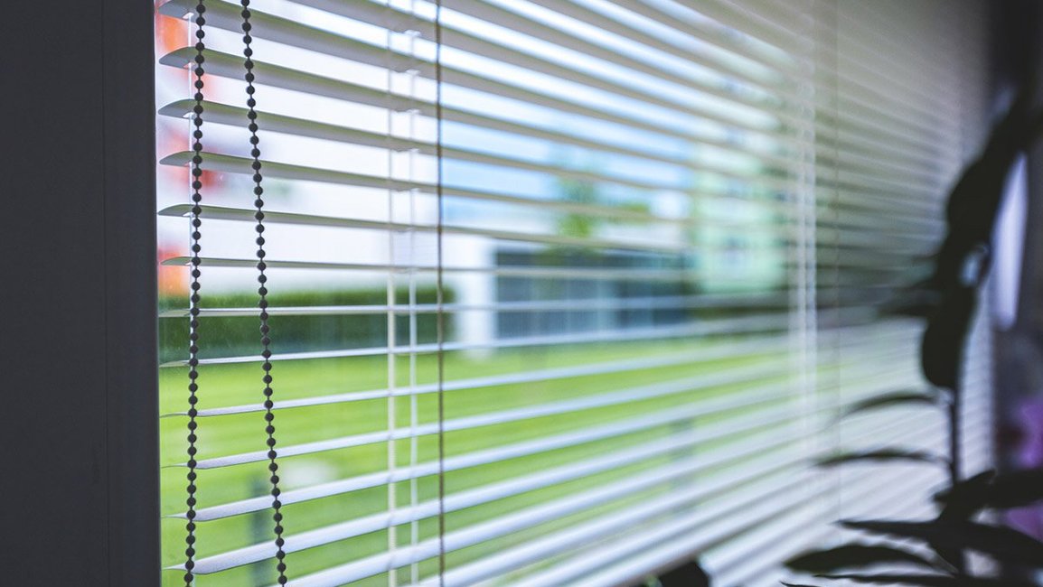 Detail bílých horizontálních žaluzií na okně rodinného domu. Lamely propouštějí příjemně rozptýlené denní světlo a zároveň zajišťují soukromí, přičemž výhled do zahrady zůstává zachovaný.
