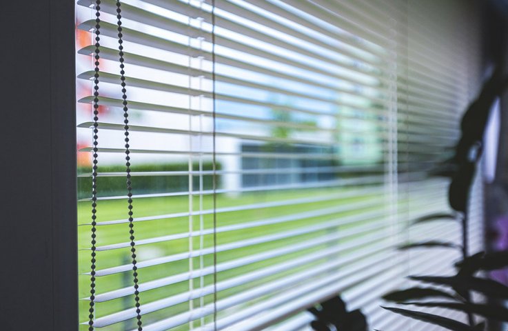 Detail bílých horizontálních žaluzií na okně rodinného domu. Lamely propouštějí příjemně rozptýlené denní světlo a zároveň zajišťují soukromí, přičemž výhled do zahrady zůstává zachovaný.