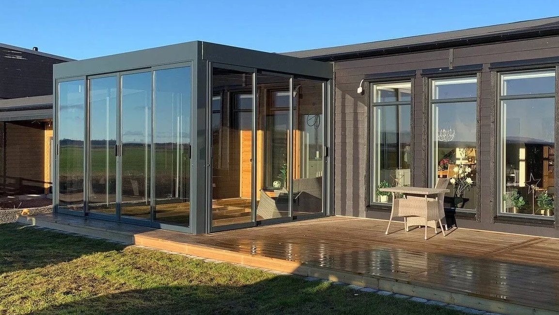 Zimní zahrada v moderní hliníkové konstrukci s velkými posuvnými skleněnými plochami plynule navazuje na dřevěnou terasu a obývací část rodinného domu. Díky celoproskleným stěnám nabízí celoročně světlý prostor s výhledem do otevřené krajiny.