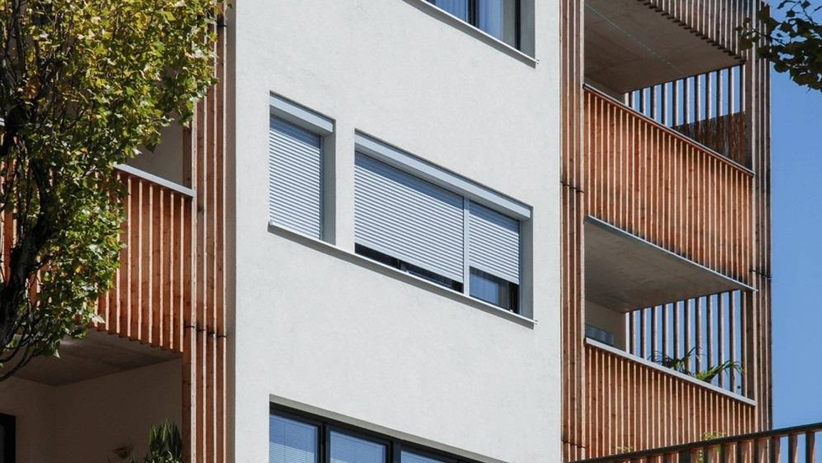 Moderní bytový dům s předokenními roletami v bílé barvě; elegantní kombinace s dřevěným lamelovým obkladem lodžií pro účinné stínění a soukromí.