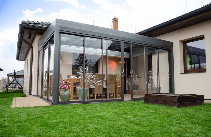 Moderní hliníková pergola u rodinného domu se zasklením po celém obvodu slouží jako komfortní venkovní jídelna. Uvnitř je velký stůl s pohodlnými židlemi, dekorace v květináčích a sálavé topení, takže si majitelé mohou užívat stolování na terase od jara až do podzimu.