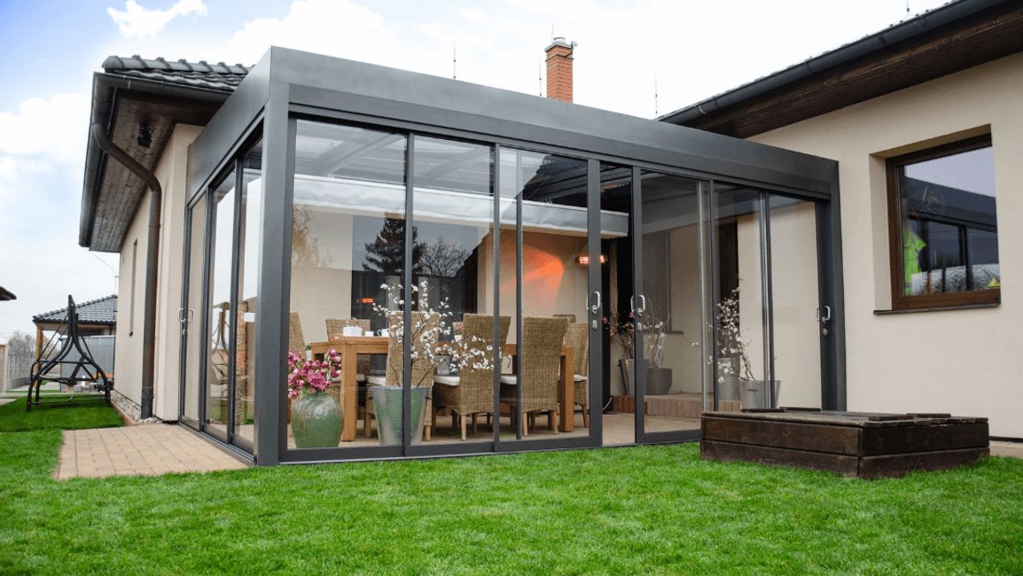 Moderní hliníková pergola u rodinného domu se zasklením po celém obvodu slouží jako komfortní venkovní jídelna. Uvnitř je velký stůl s pohodlnými židlemi, dekorace v květináčích a sálavé topení, takže si majitelé mohou užívat stolování na terase od jara až do podzimu.