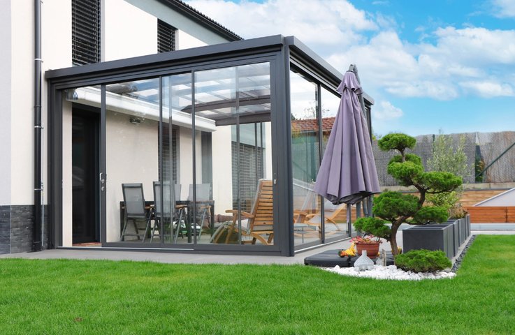 Moderní hliníková pergola u rodinného domu je zasklená posuvnými skleněnými stěnami a se skleněnou střechou vytváří chráněnou terasu pro celoroční posezení.