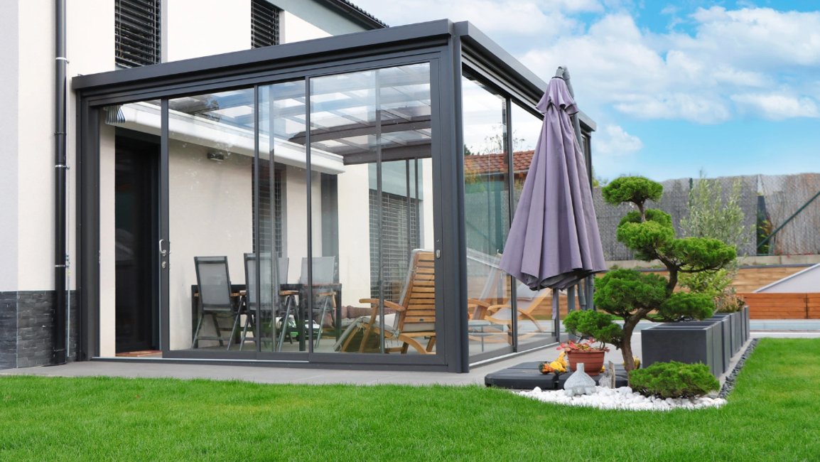 Moderní hliníková pergola u rodinného domu je zasklená posuvnými skleněnými stěnami a se skleněnou střechou vytváří chráněnou terasu pro celoroční posezení.