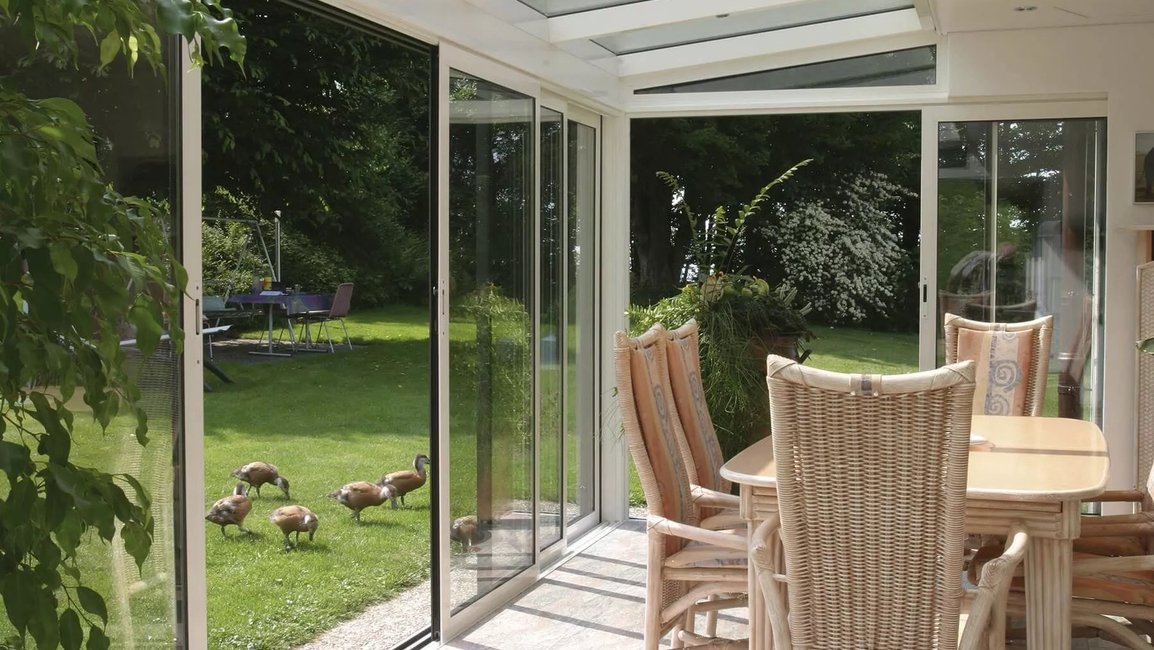 Jídelní kout v zimní zahradě je chráněný rámovým posuvným zasklením a prosklenou střechou. Velké skleněné plochy zajišťují dostatek denního světla a umožňují pohodlný vstup přímo na trávník, takže se interiér přirozeně propojuje se zahradou.