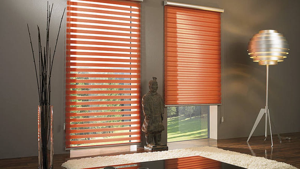 Rolety den a noc v oranžové barvě v moderním obývacím pokoji – jedno okno v režimu průhledných pruhů, druhé v plném zatemnění.