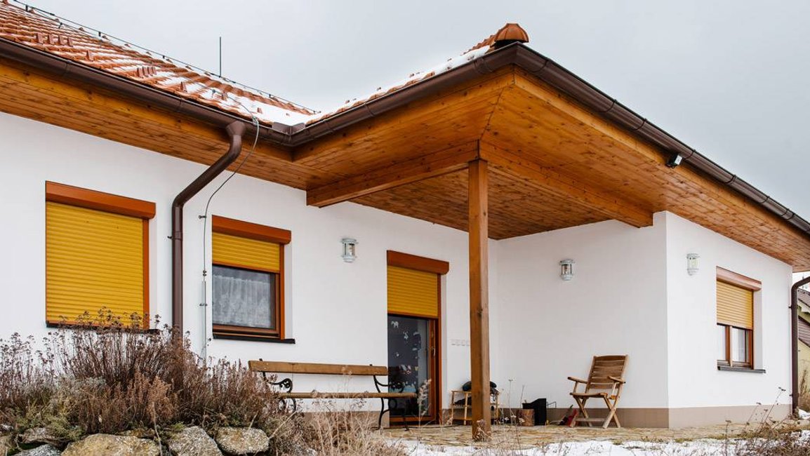 Moderní rodinný dům s bílo–dřevěnou fasádou a žlutými venkovními předokenními roletami, které chrání okna před sluncem a zároveň ladí s přírodním okolím.
