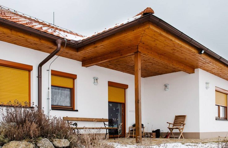 Moderní rodinný dům s bílo–dřevěnou fasádou a žlutými venkovními předokenními roletami, které chrání okna před sluncem a zároveň ladí s přírodním okolím.